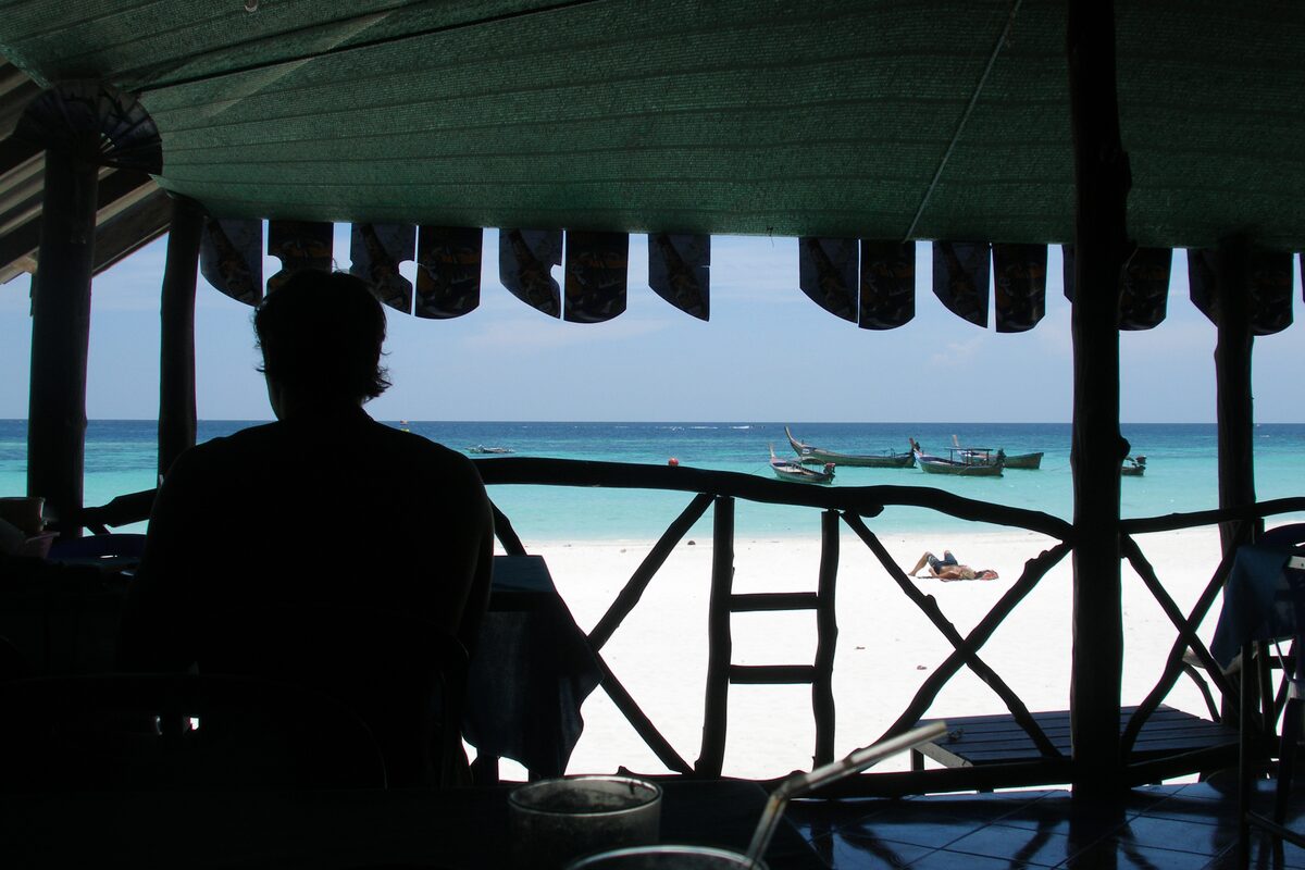 View from a simple beach hut: a guest in silhouette, white sand and longtail boats on turquoise water