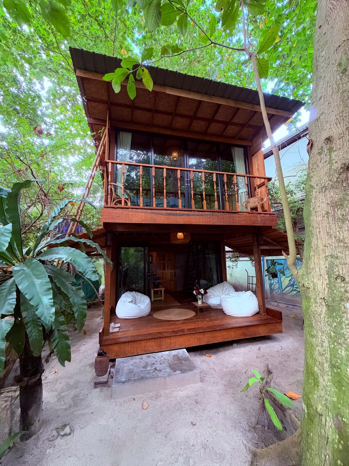 Wooden bungalow seen from the sandy garden, with a balcony and a covered veranda