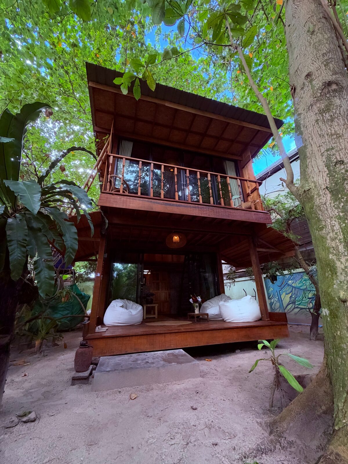 Two-storey wooden bungalow under tall trees: balcony upstairs, veranda with white beanbags below